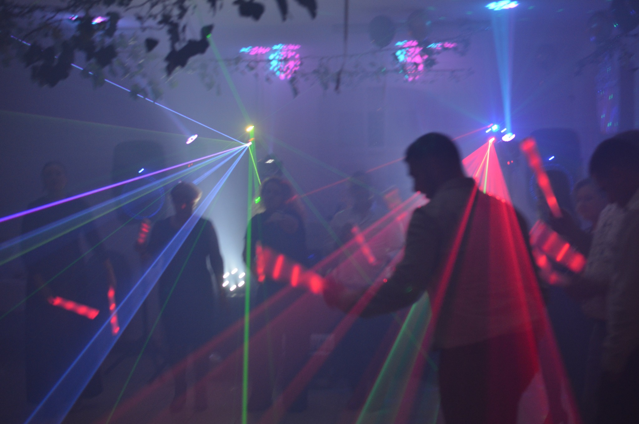 Impreza w klubie z efektami laserowymi: zielone, niebieskie i czerwone promienie przecinają przestrzeń, oświetlając tańczących ludzi w tle. Dynamiczne oświetlenie.
