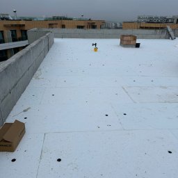 Kam-Dach - Białe płyty izolacyjne na płaskim dachu budynku wielorodzinnego, widoczne narzędzia i materiały budowlane. Szary, pochmurny dzień.