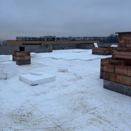 Kam-Dach - Ocieplenie płaskiego dachu styropianem, widoczne ceglane kominy i odległe budynki na horyzoncie, pochmurne niebo. Prace dekarskie w toku.