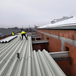 Kam-Dach - Montaż blachy trapezowej na dachu budynku w trakcie budowy. Widoczni dekarze w odblaskowych kamizelkach i elementy konstrukcyjne dachu.