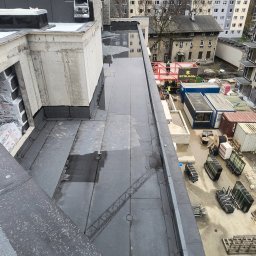 Kam-Dach - Wykończony, płaski dach budynku z papy termozgrzewalnej, widok z góry. Na dachu widoczne kałuże. W tle budynki mieszkalne i kontenery budowlane.