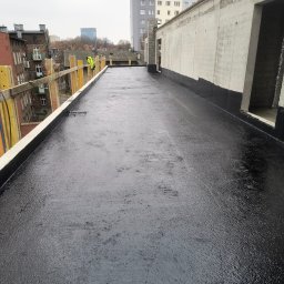 Kam-Dach - Czarna, płaska powierzchnia dachu po hydroizolacji, z widocznymi elementami konstrukcyjnymi i fragmentem miasta w tle. Prace dekarskie w toku.