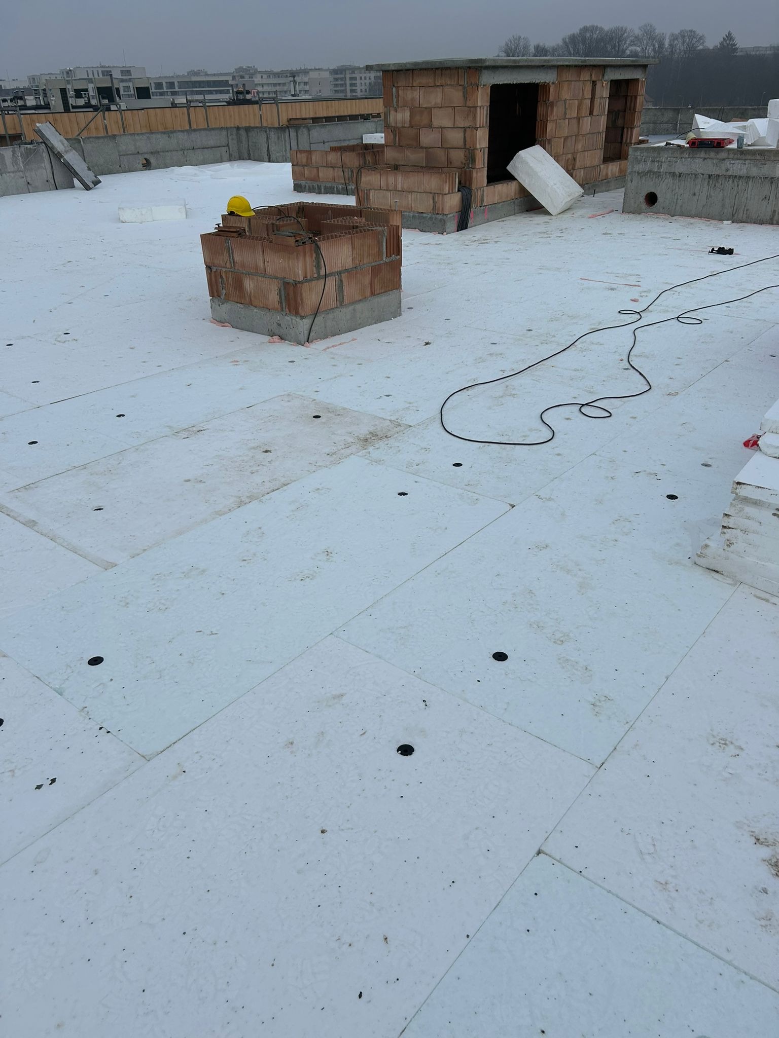 Białe płyty izolacyjne na dachu budynku w trakcie budowy, widoczne ceglane konstrukcje i żółty kask. Przewody elektryczne na powierzchni izolacji.