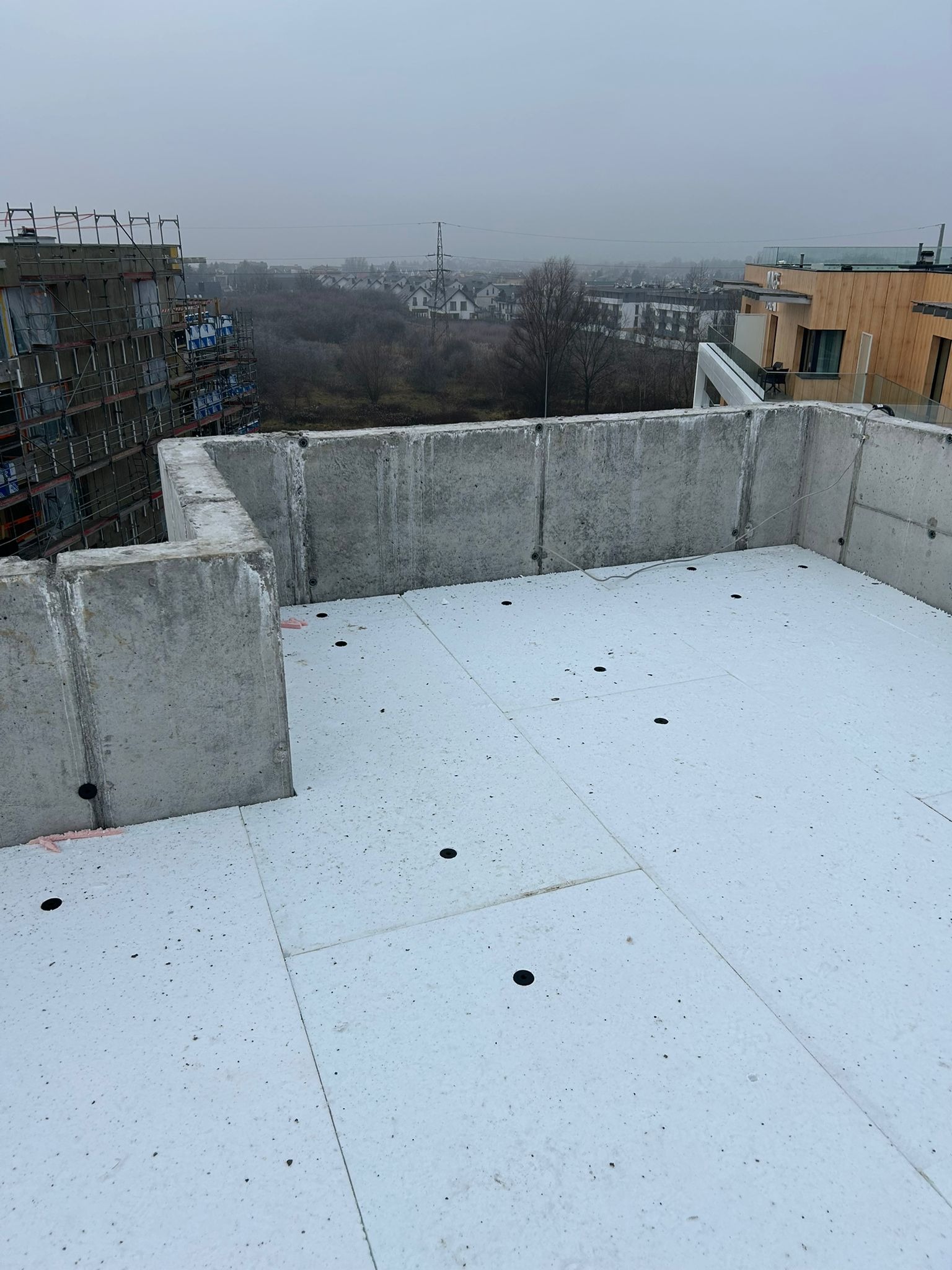 Betonowa konstrukcja dachu płaskiego z izolacją termiczną, widok z góry na tle zabudowy miejskiej w pochmurny dzień. Widoczne otwory w izolacji.