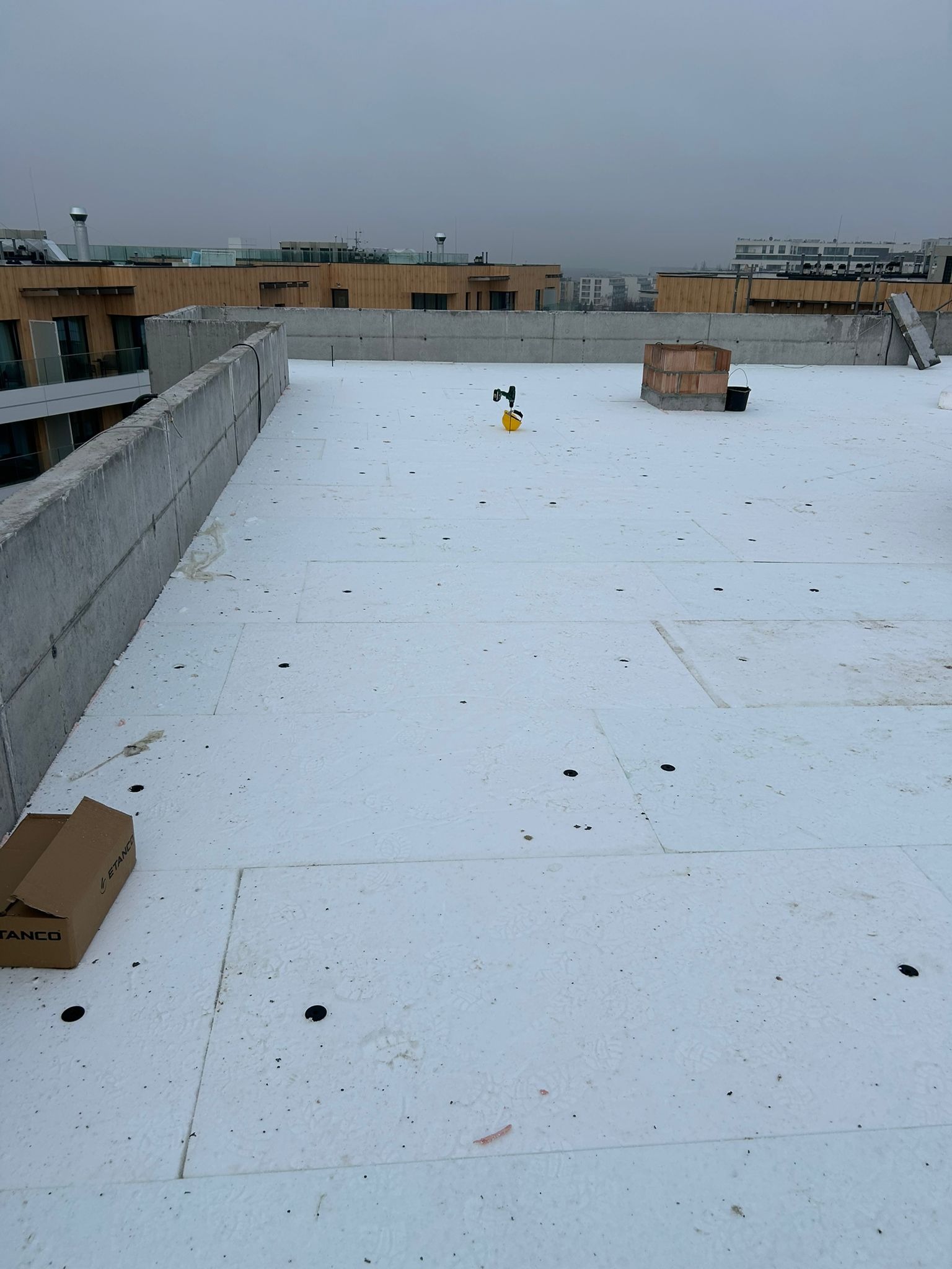 Białe płyty izolacyjne na płaskim dachu budynku wielorodzinnego, widoczne narzędzia i materiały budowlane. Szary, pochmurny dzień.