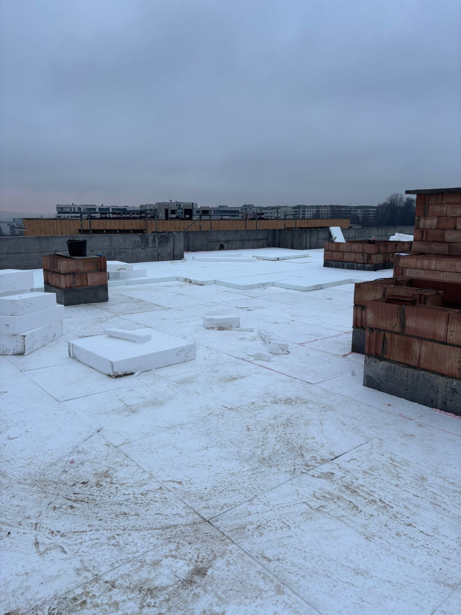 Ocieplenie płaskiego dachu styropianem, widoczne ceglane kominy i odległe budynki na horyzoncie, pochmurne niebo. Prace dekarskie w toku.