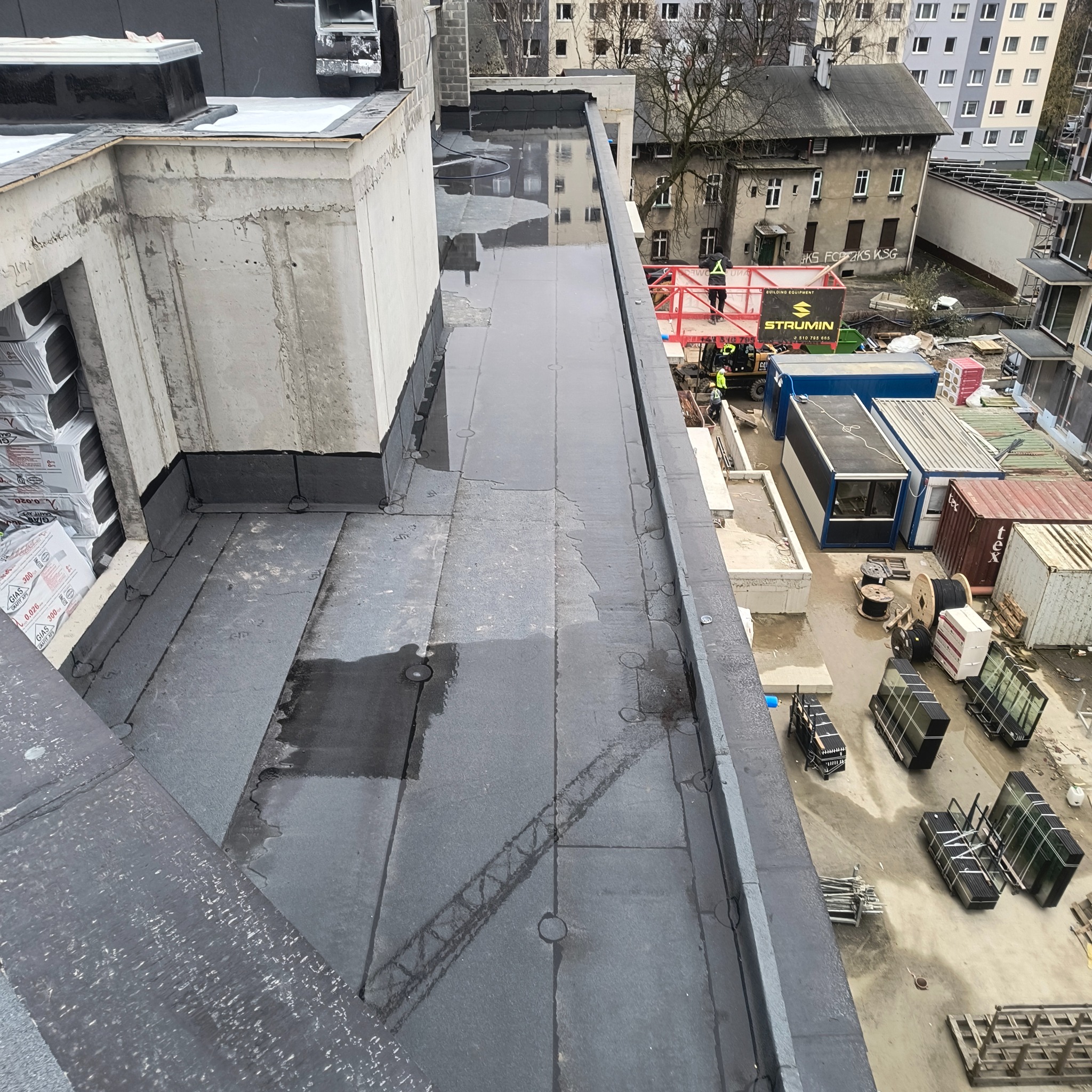 Wykończony, płaski dach budynku z papy termozgrzewalnej, widok z góry. Na dachu widoczne kałuże. W tle budynki mieszkalne i kontenery budowlane.