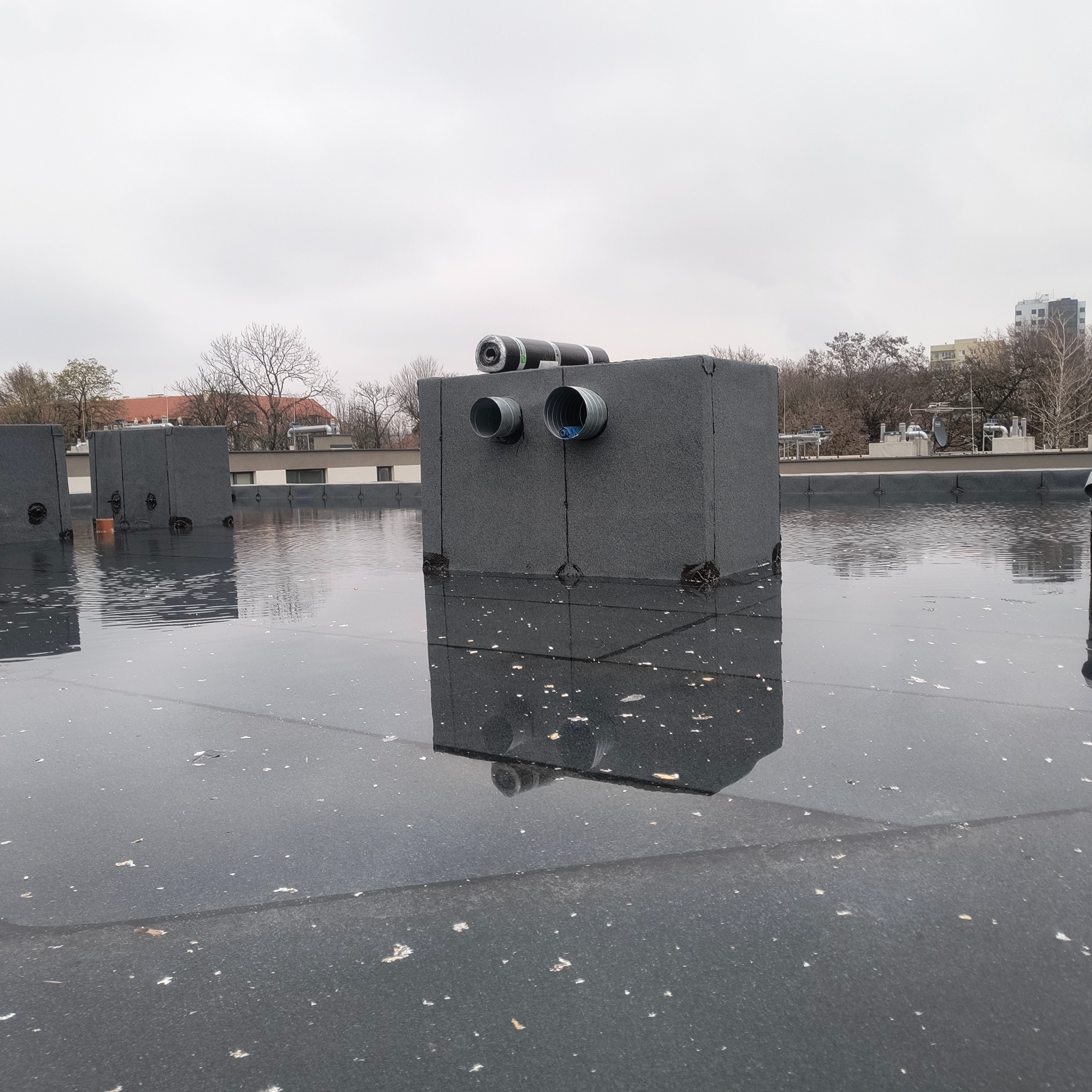 Płaski dach pokryty papą z odpływami i odpowietrznikami, częściowo zalany wodą po deszczu. Widoczne odbicia w wodzie i budynki w tle.