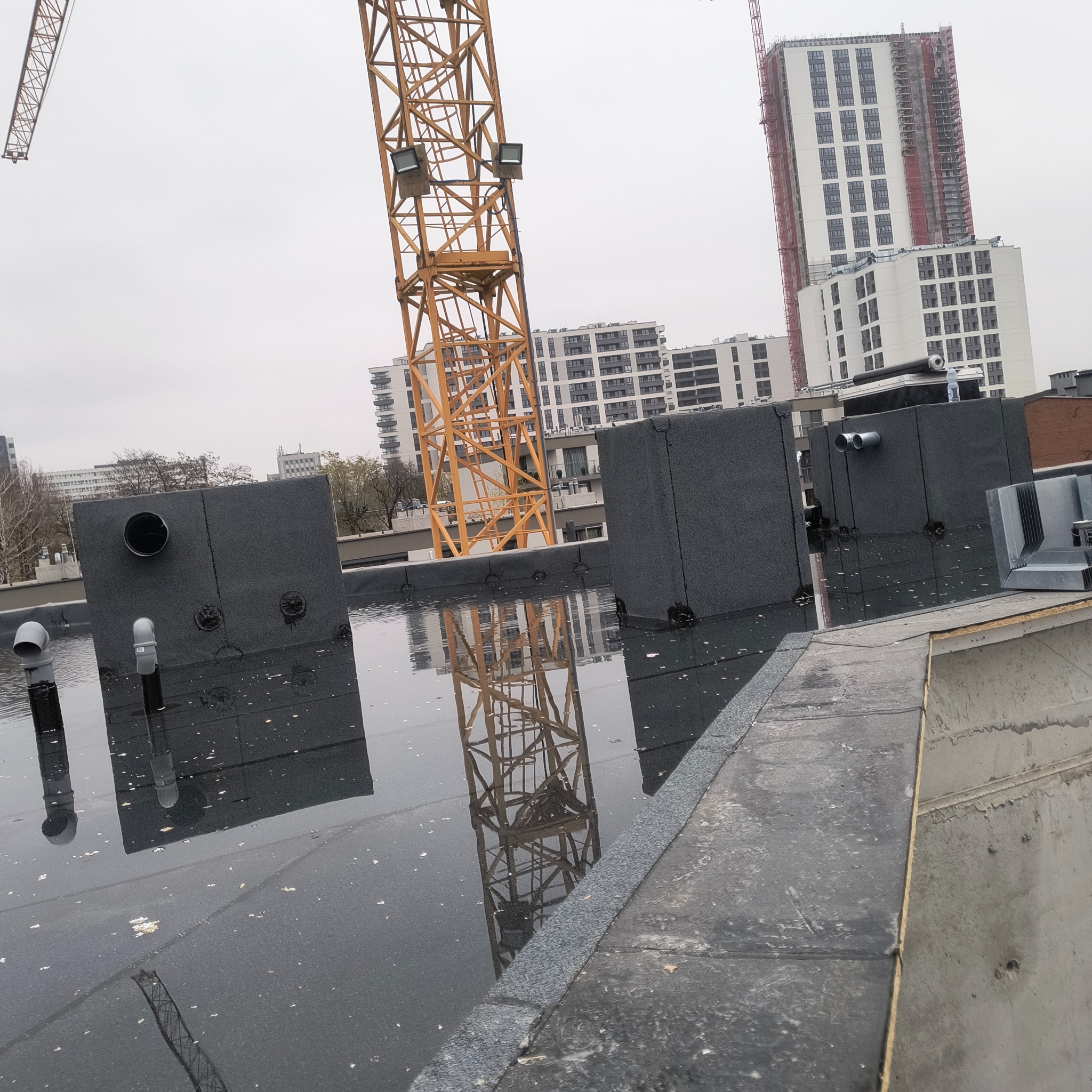 Płaski dach pokryty papą z widocznymi elementami wentylacyjnymi i odbiciem żurawia w kałużach. W tle budynki w trakcie budowy.
