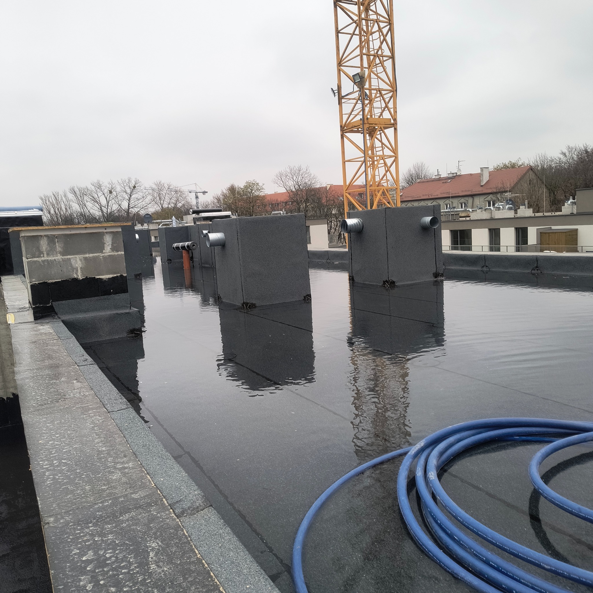Wodoodporna izolacja dachu płaskiego z elementami wentylacyjnymi i niebieskim wężem na powierzchni. W tle widoczny żuraw budowlany i budynki.