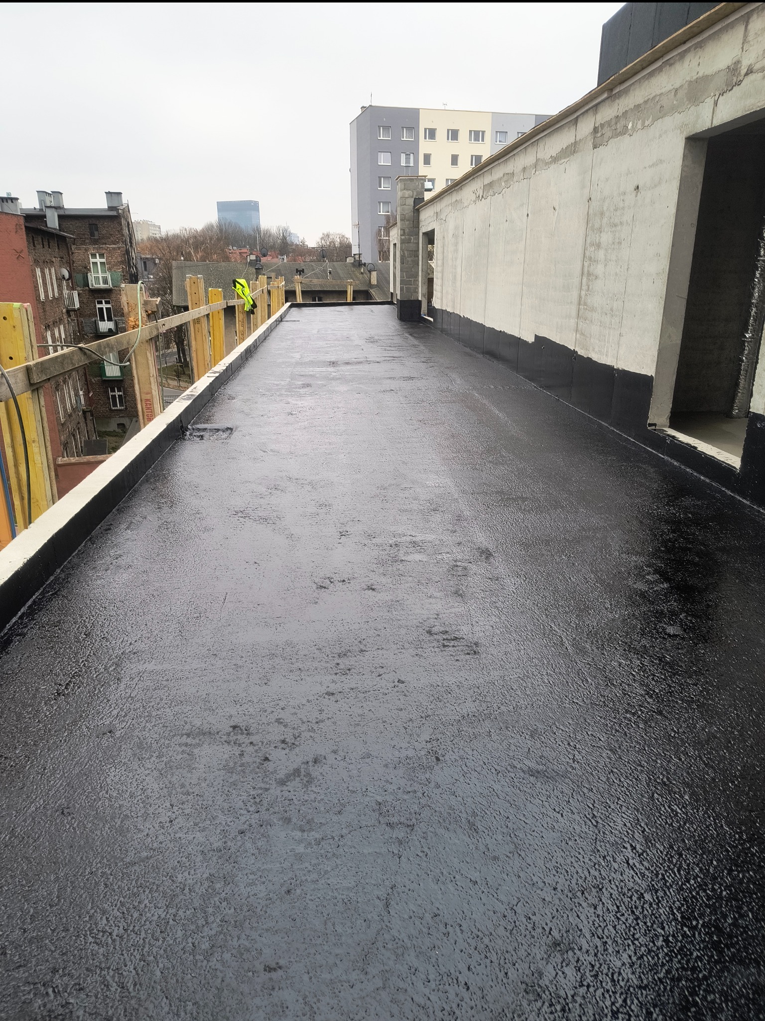 Czarna, płaska powierzchnia dachu po hydroizolacji, z widocznymi elementami konstrukcyjnymi i fragmentem miasta w tle. Prace dekarskie w toku.