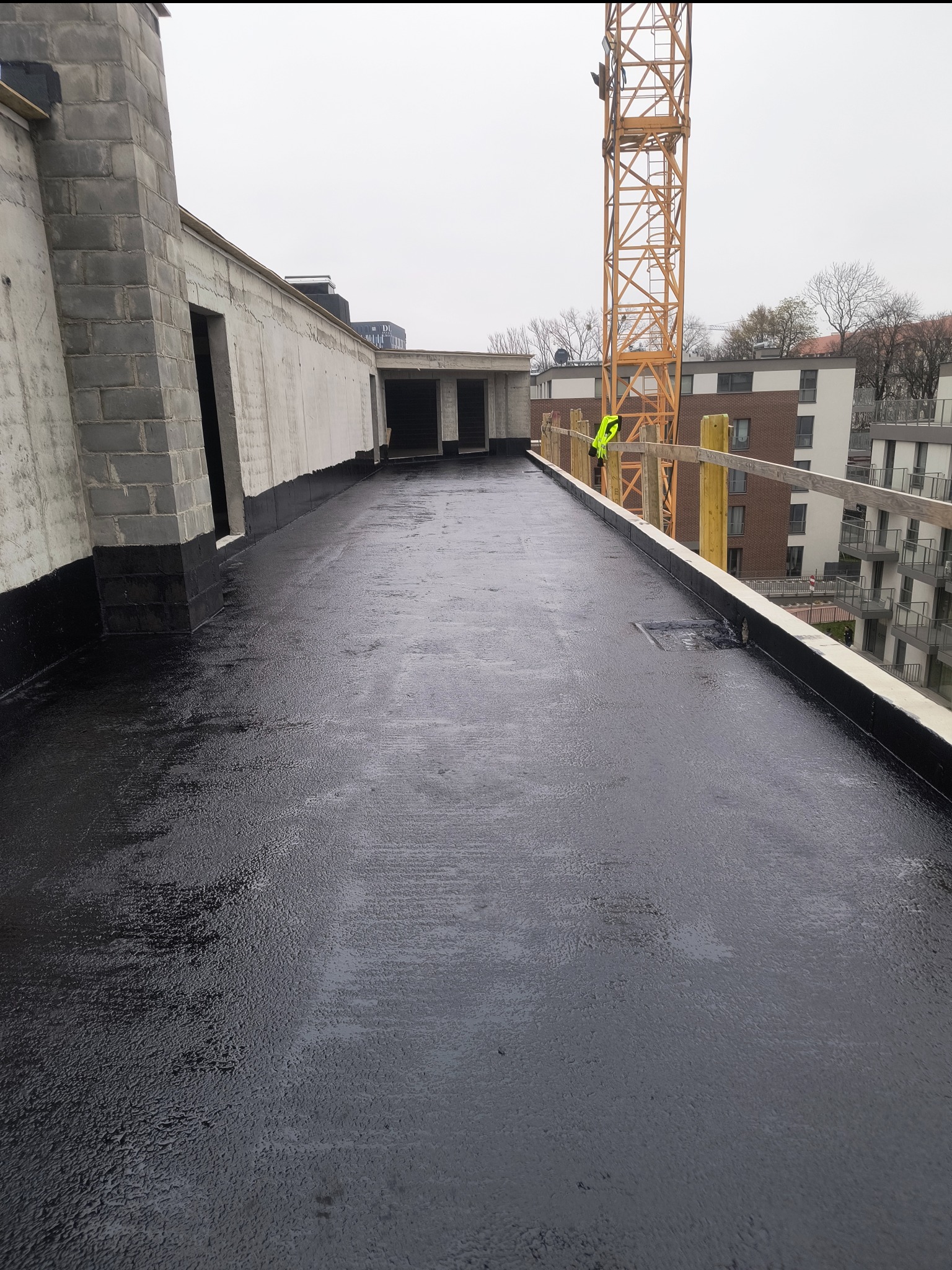 Czarna, płaska powierzchnia dachu po hydroizolacji, z widocznym fragmentem budynku w stanie surowym i żurawiem budowlanym w tle. Praca dekarska.