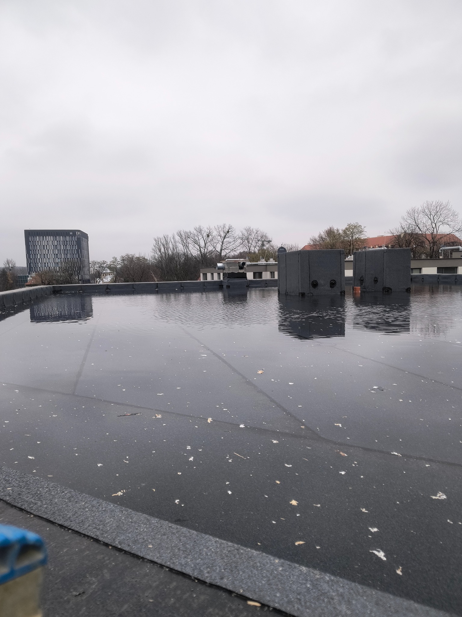 Płaski dach pokryty wodą po deszczu, z widocznymi elementami konstrukcyjnymi i budynkami w tle, pochmurne niebo. Widoczne zabrudzenia na powierzchni dachu.