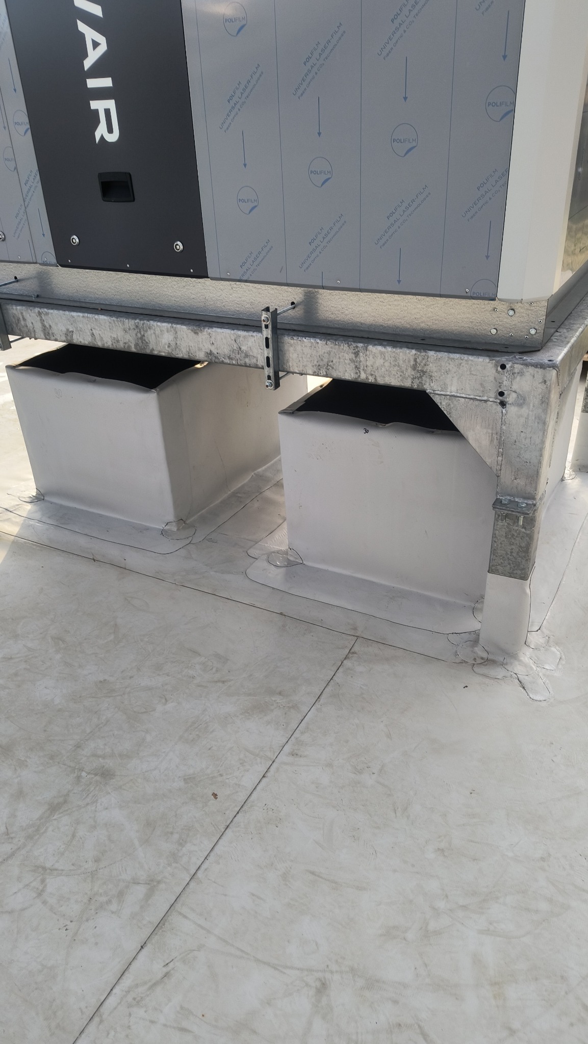 Szczelne przejścia wentylacyjne na dachu pokrytym membraną, zabezpieczone białą masą uszczelniającą. Widoczne elementy konstrukcji wsporczej i fragment urządzenia wentylacyjnego.