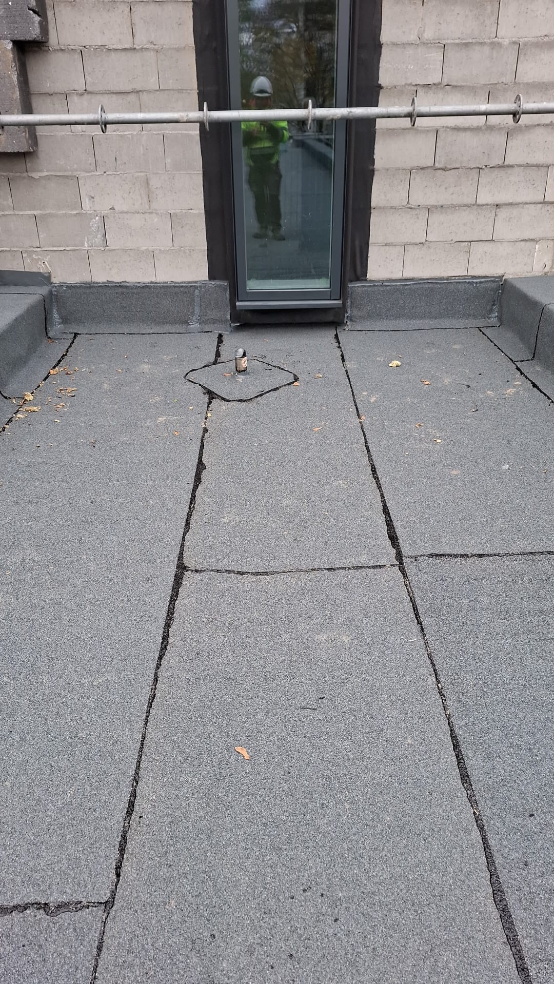 Szczegółowe ujęcie płaskiego dachu pokrytego papą, z widocznym oknem dachowym i elementami mocującymi. Widoczna faktura papy i łączenia. W tle mur z bloczków.