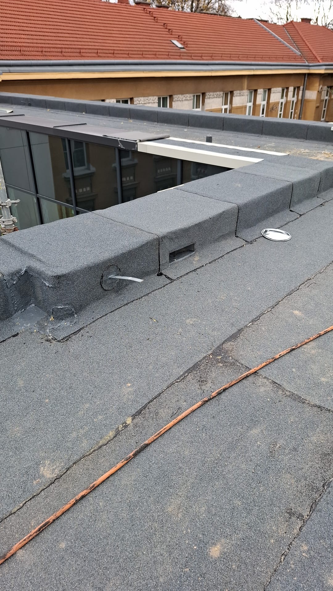 Szary, płaski dach pokryty papą z widocznymi elementami wentylacji i odpływem. W tle budynek z czerwoną dachówką i oknami.