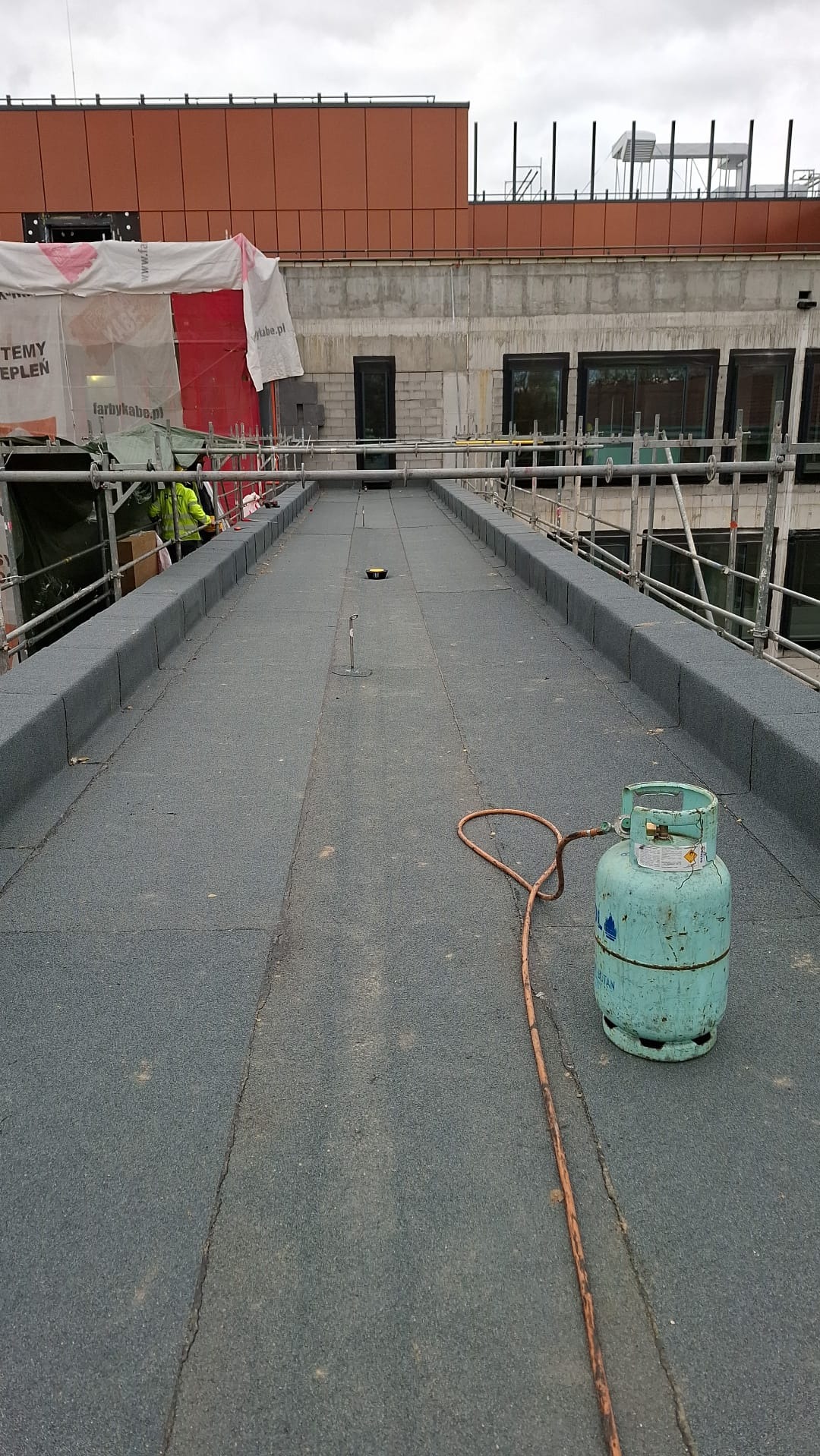 Szary dach płaski z widocznym zbiornikiem gazu i fragmentem budynku w tle. Prace dekarskie w toku, widoczne rusztowania i materiały budowlane.