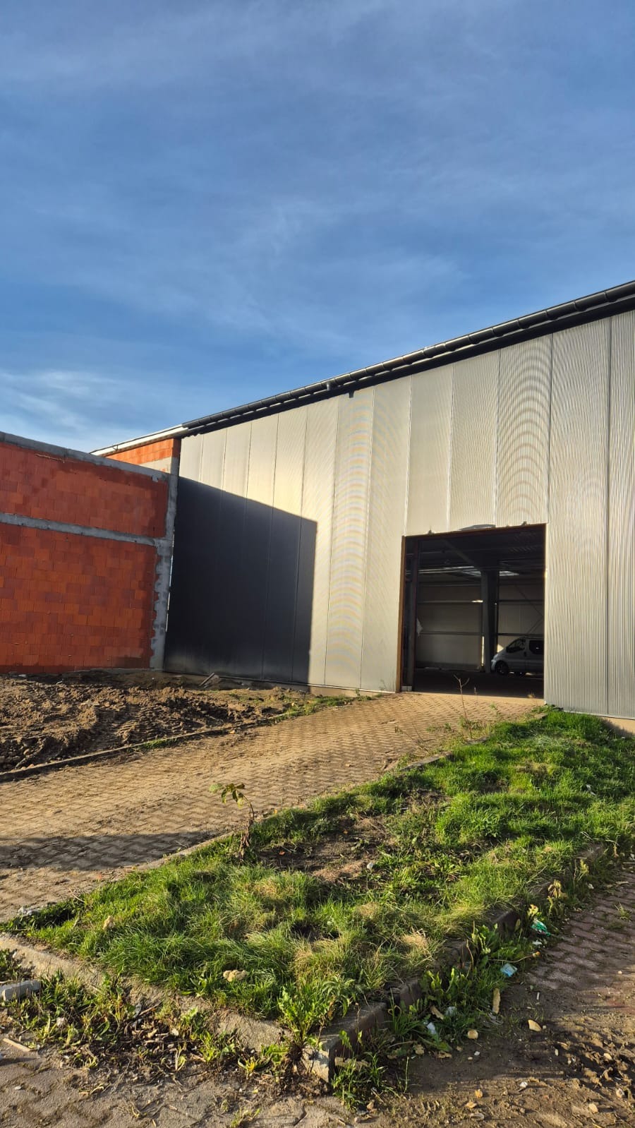 Budynek w trakcie budowy z blachy trapezowej i cegły, widoczny fragment dachu i brama wjazdowa. Teren wokół budynku częściowo zagospodarowany zielenią.