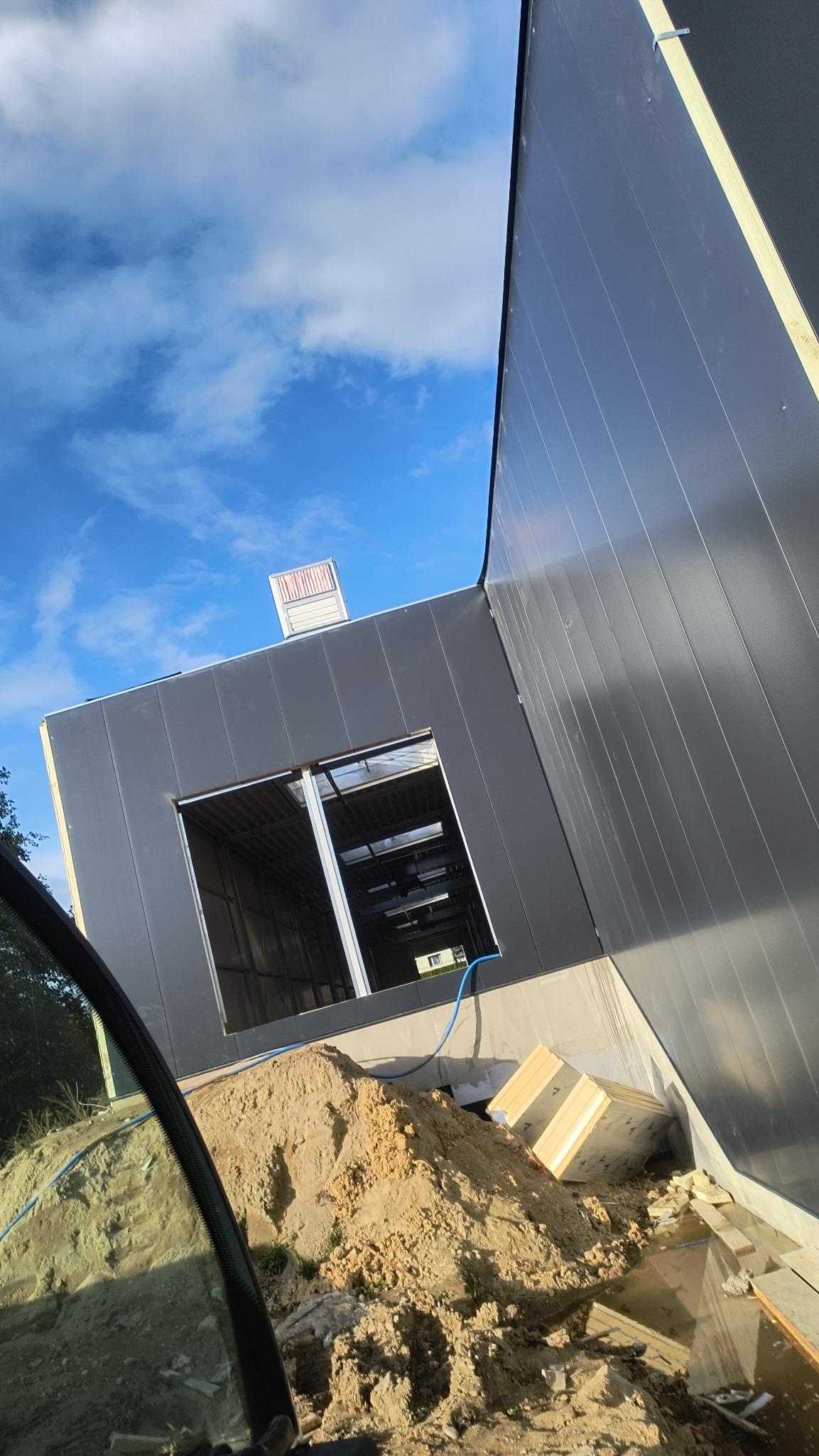 Budynek w trakcie budowy z szarej blachy trapezowej, widok z dołu na niebo. Widoczne okno bez szyby, piasek i materiały budowlane. Ujęcie zza szyby pojazdu.