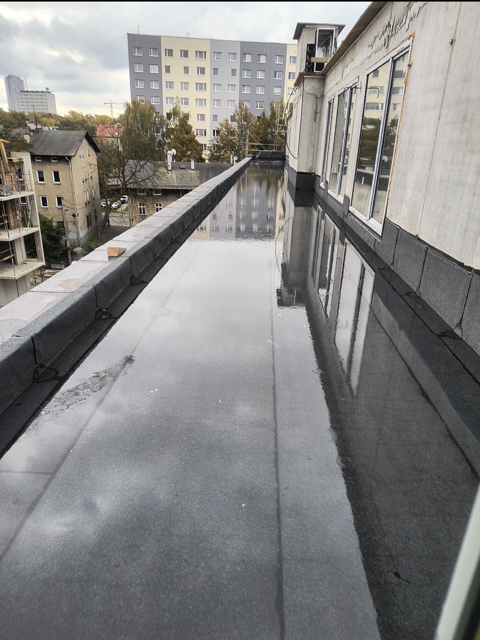 Wodoodporna, czarna papa na płaskim dachu budynku po deszczu. Widoczne okna i budynki w tle. Detal hydroizolacji dachu z papy termozgrzewalnej.