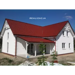 Biały dom jednorodzinny z czerwonym dachem, widok z zewnątrz, częściowo zagospodarowany teren, widoczne materiały budowlane i butelki z wodą.