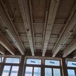 Michał Kliś - Drewniany strop z widocznymi belkami i instalacją elektryczną, surowy stan wykończenia, duże okna w tle. Widok z dołu.