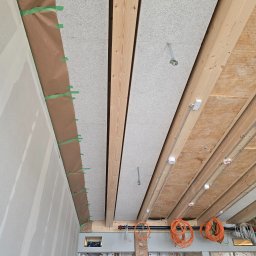 Michał Kliś - Montaż paneli akustycznych na drewnianej konstrukcji sufitu, widoczne przewody elektryczne i fragment ściany z płyt kartonowo-gipsowych zabezpieczonej folią malarską.