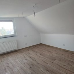 Michał Kliś - Poddasze po remoncie: drewnopodobna podłoga, białe ściany, okno z grzejnikiem. Widoczne gniazdka i wiszące kable. Minimalistyczny design, skosy sufitu.