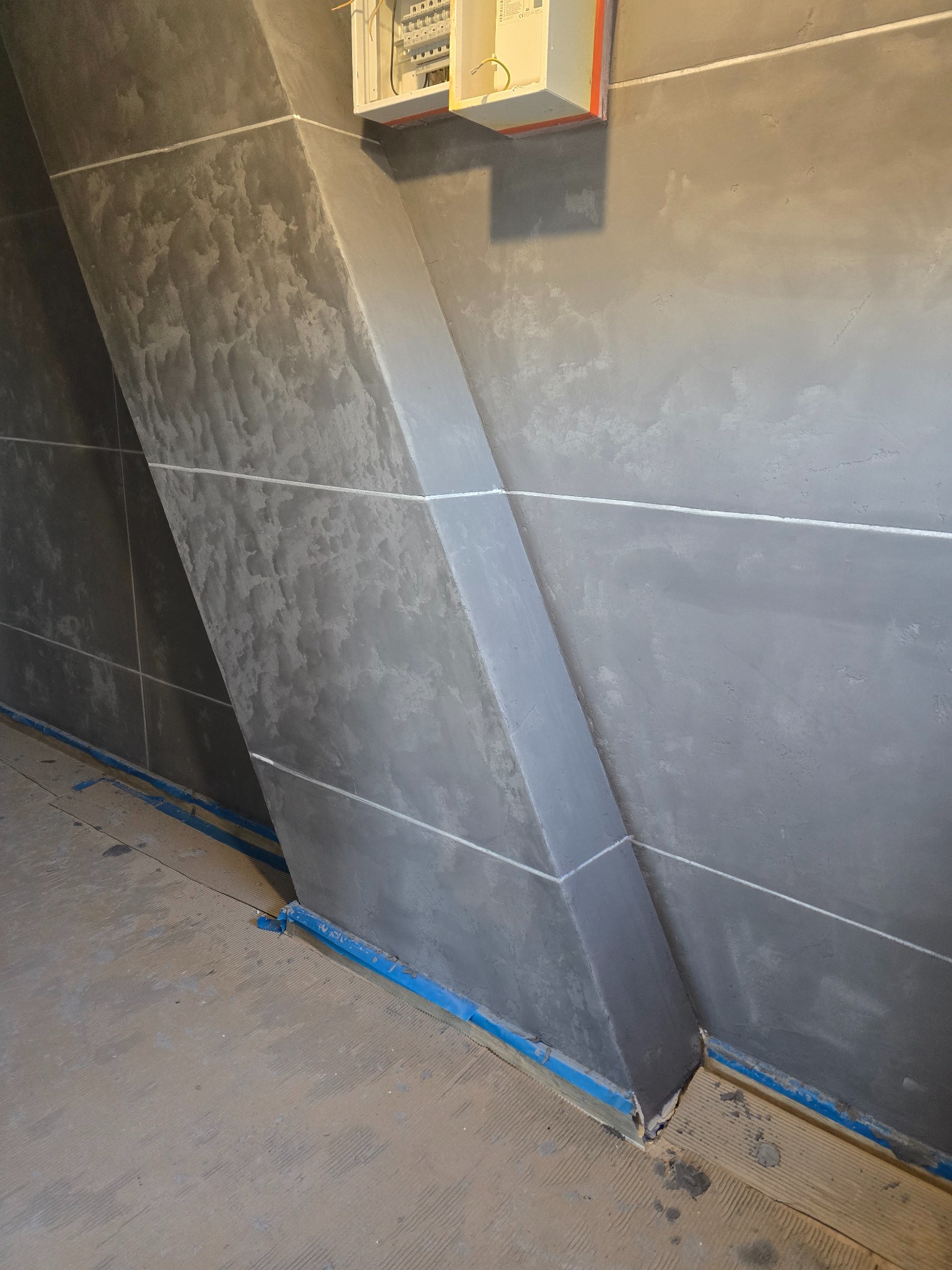 Szara ściana z dekoracyjną strukturą i wystającym elementem architektonicznym, zabezpieczona niebieską taśmą malarską przy podłodze. Widoczna skrzynka elektryczna.