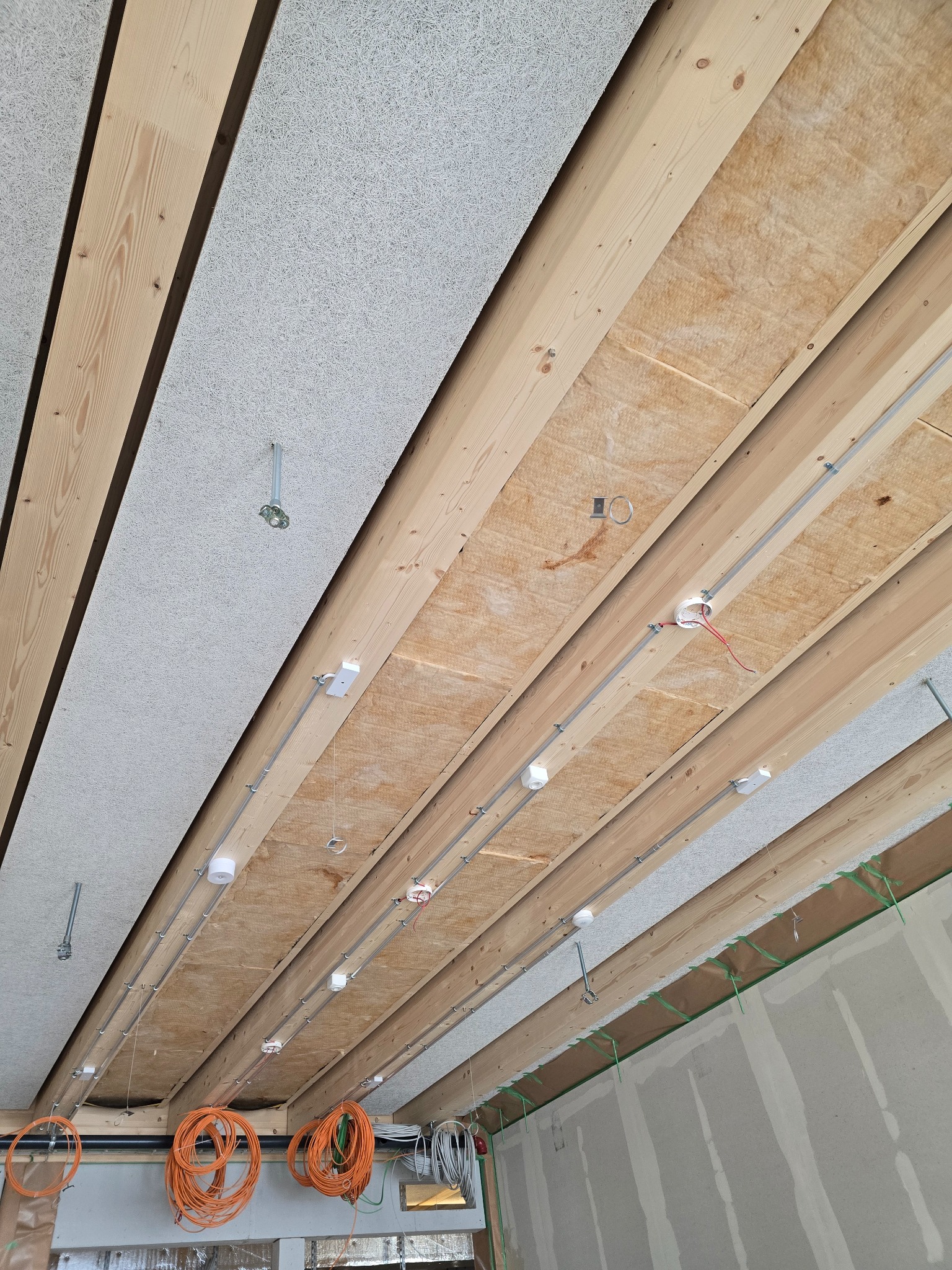 Drewniana konstrukcja sufitu z izolacją, instalacją elektryczną i panelami akustycznymi. Widoczne kable i elementy montażowe. Ujęcie z dołu, perspektywa zbieżna.