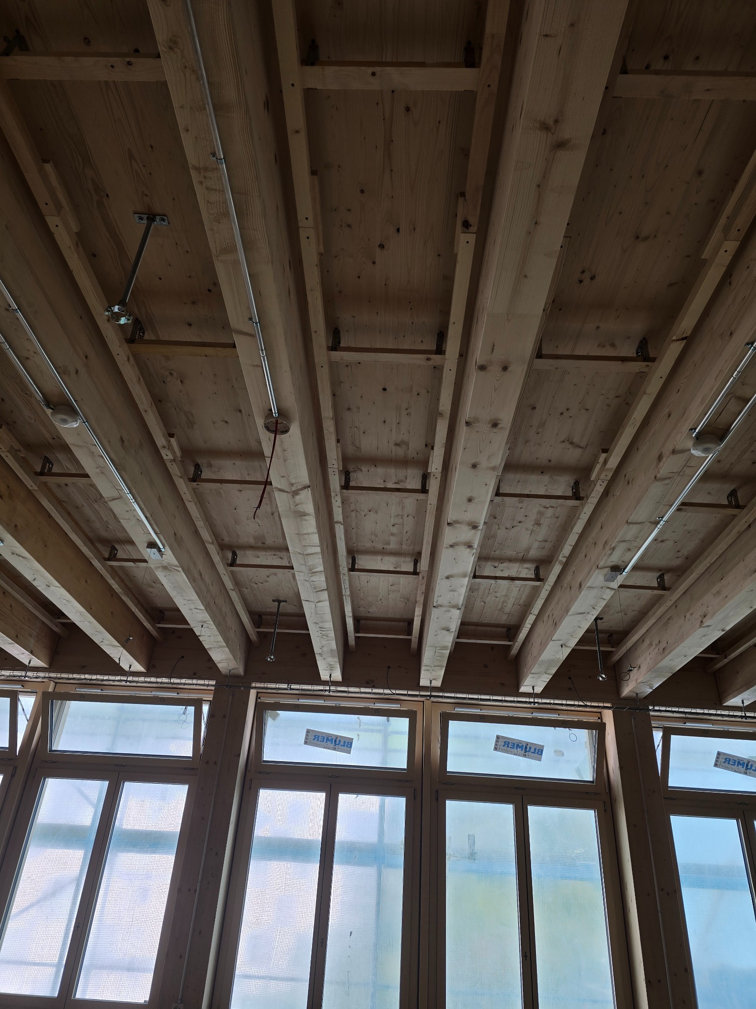 Drewniany strop z widocznymi belkami i instalacją elektryczną, surowy stan wykończenia, duże okna w tle. Widok z dołu.