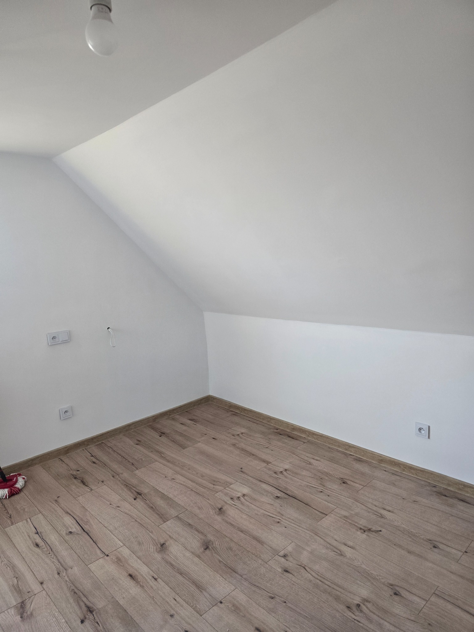 Poddasze po remoncie: świeżo ułożona podłoga z paneli, białe ściany ze skosami, gniazdka elektryczne. Widoczny fragment mopa w rogu.