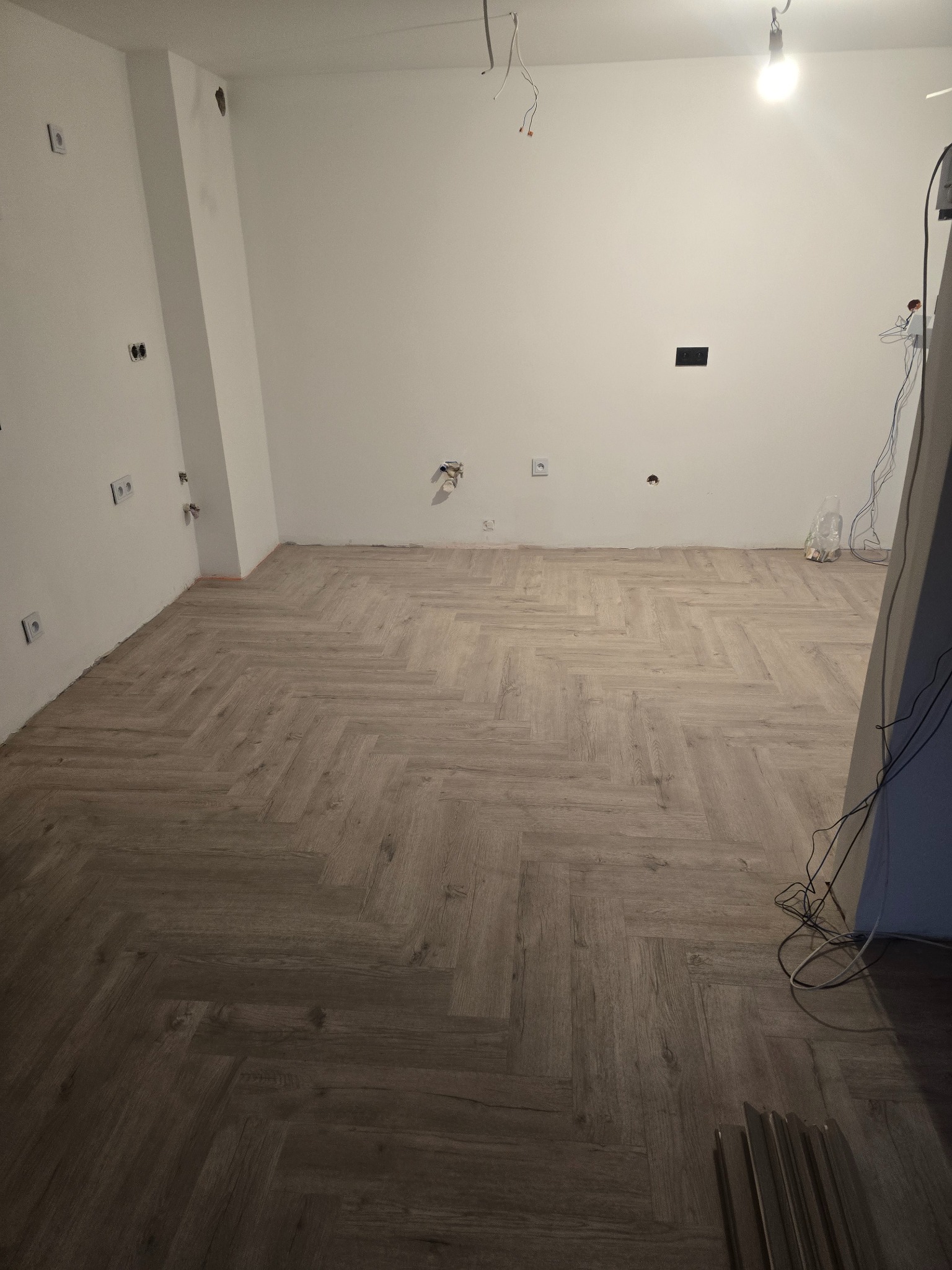 Jasny parkiet ułożony w jodełkę w pustym pokoju z białymi ścianami i widocznymi gniazdkami elektrycznymi. W tle widoczne kable i niedokończone wykończenia.