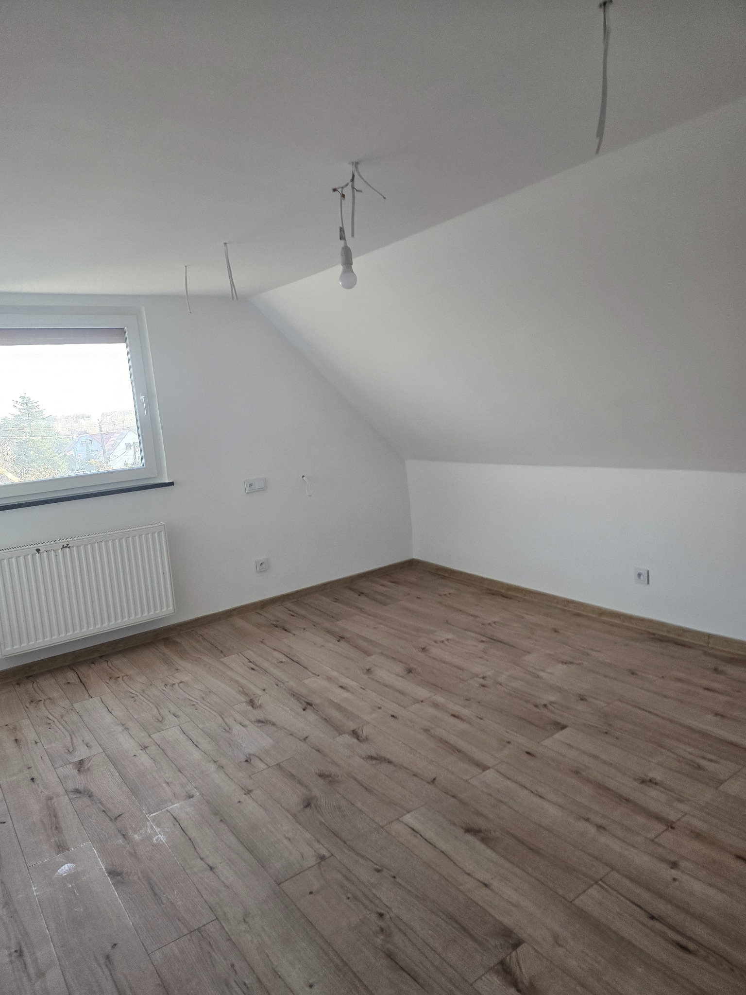 Poddasze po remoncie: drewnopodobna podłoga, białe ściany, okno z grzejnikiem. Widoczne gniazdka i wiszące kable. Minimalistyczny design, skosy sufitu.