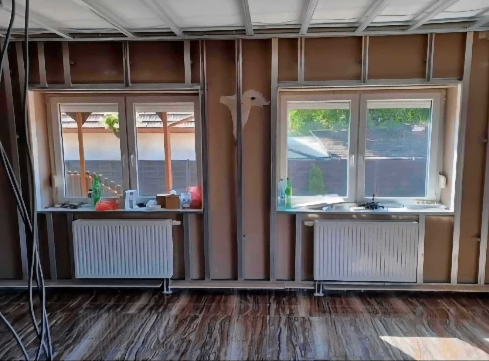 Wnętrze w trakcie prac wykończeniowych: widoczne profile pod płyty gipsowe, okna z grzejnikami, podłoga imitująca drewno. Częściowo zaszpachlowana ściana.