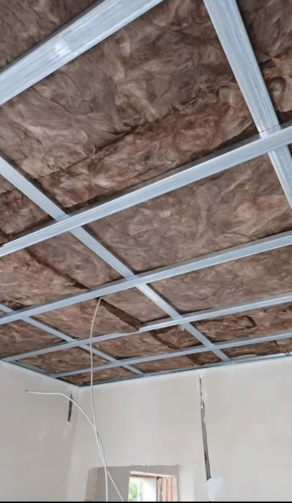 Metalowy stelaż sufitowy z izolacją z wełny mineralnej, widok z dołu, przewody elektryczne zwisają. Etap prac wykończeniowych wnętrza.