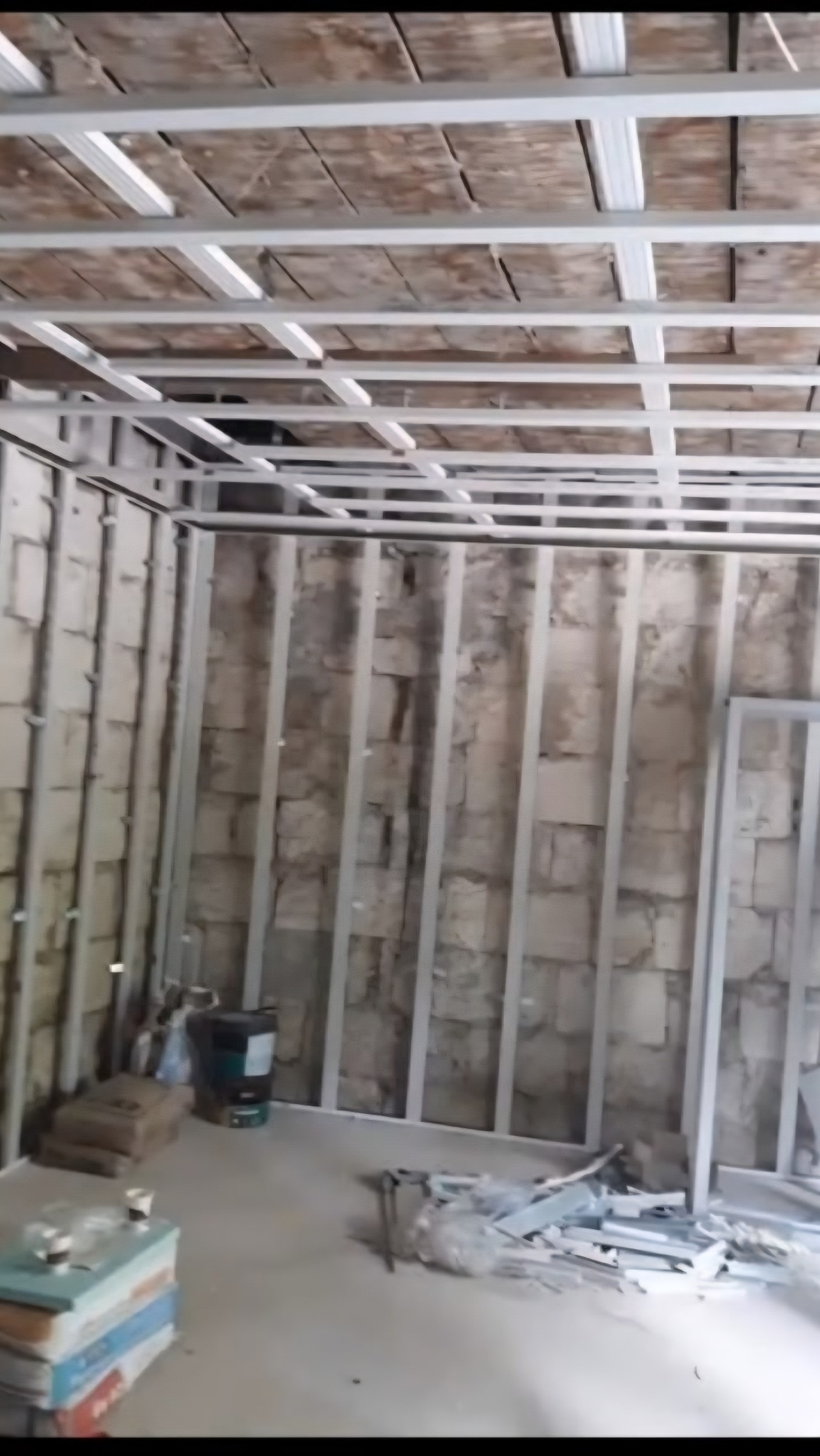 Montaż stelaża sufitu podwieszanego z płyt karton-gips, widoczne profile metalowe i fragment starego sufitu. Na podłodze materiały budowlane i narzędzia.