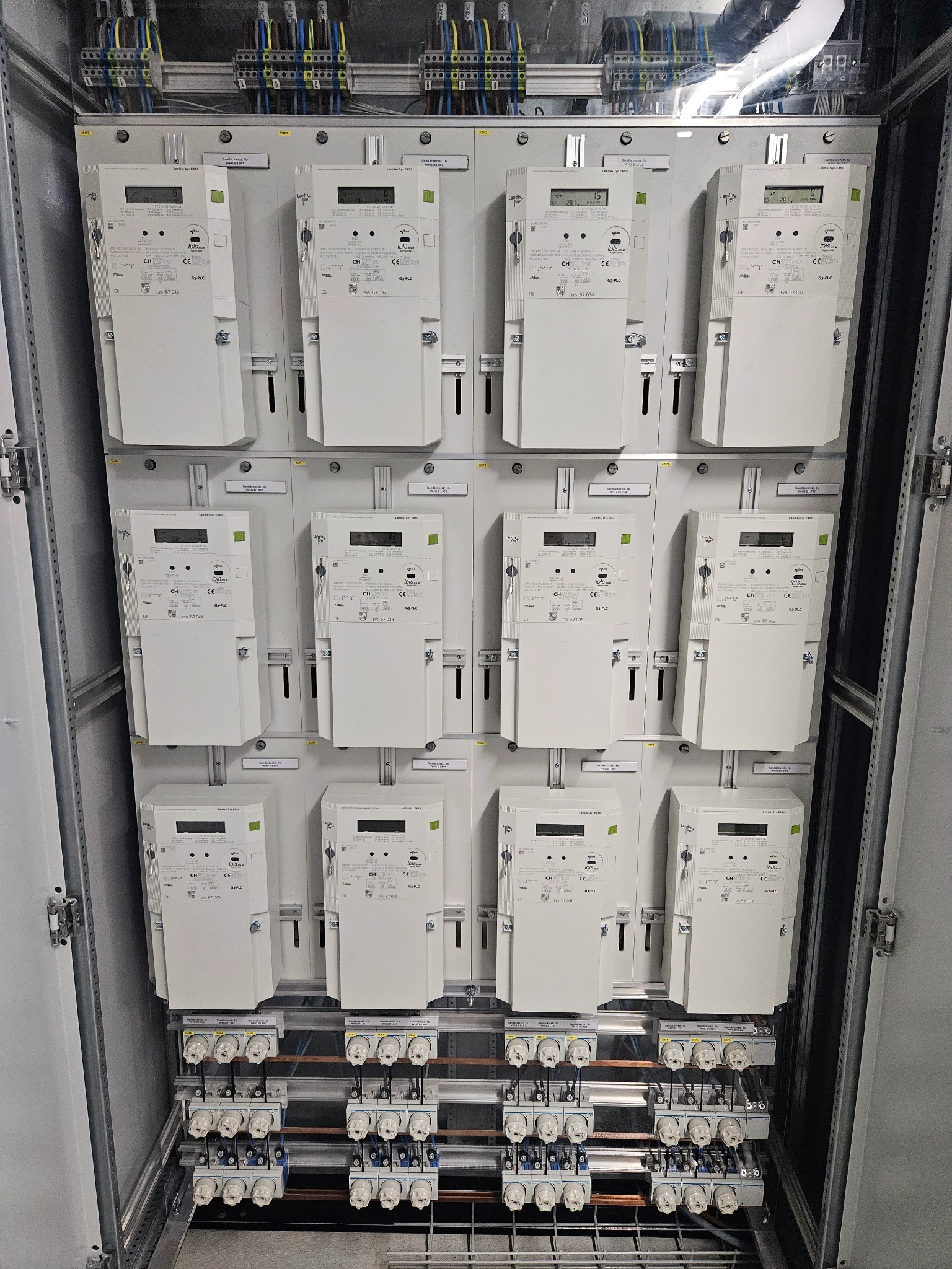 Rozdzielnica elektryczna z zamontowanymi licznikami energii elektrycznej, widoczne bezpieczniki i okablowanie. Precyzyjny montaż w szarej obudowie.