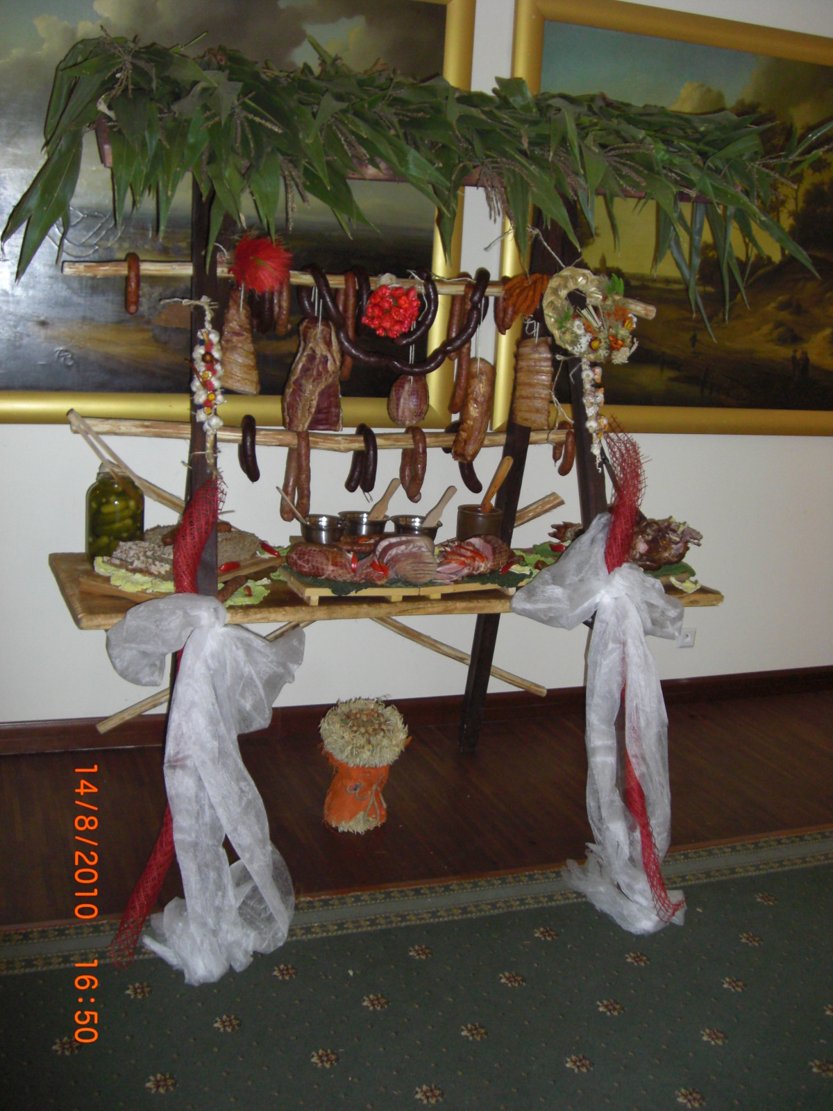 Stół wiejski z wędlinami, kiełbasami i potrawami mięsnymi, udekorowany liśćmi kukurydzy i białymi wstążkami, na tle obrazów w złotych ramach.