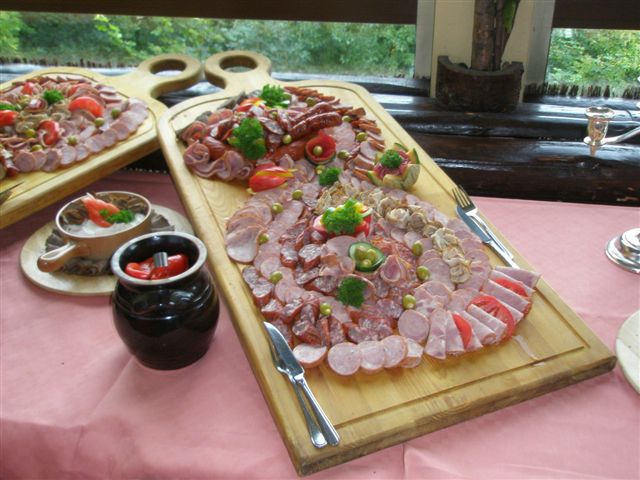 Stół wiejski z deskami pełnymi wędlin, kiełbas i dekoracji z ziół i warzyw, ustawiony na różowym obrusie.