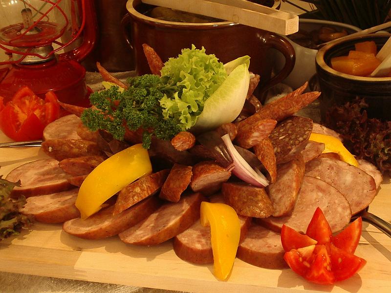 Stół z wędlinami i dekoracjami warzywnymi, w tle gliniane naczynia i czerwona lampa naftowa, ułożone na drewnianej desce.