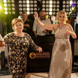 M&M EVENTS MAREK MURAWSKI - Uśmiechnięte kobiety tańczą na parkiecie, w tle DJ Marek Murawski i oświetlenie sceniczne. Radosna atmosfera imprezy.