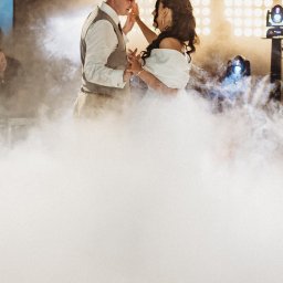 M&M EVENTS MAREK MURAWSKI - Para młoda tańczy w dymie na tle świateł scenicznych. Pan młody w kamizelce, panna młoda w białej sukni. Romantyczna atmosfera, oświetlenie eventowe.
