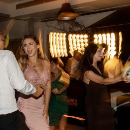 M&M EVENTS MAREK MURAWSKI - Roztańczona grupa osób na parkiecie, oświetlenie sceniczne tworzy dynamiczną atmosferę. Widoczny sprzęt nagłośnieniowy i dekoracje świetlne.