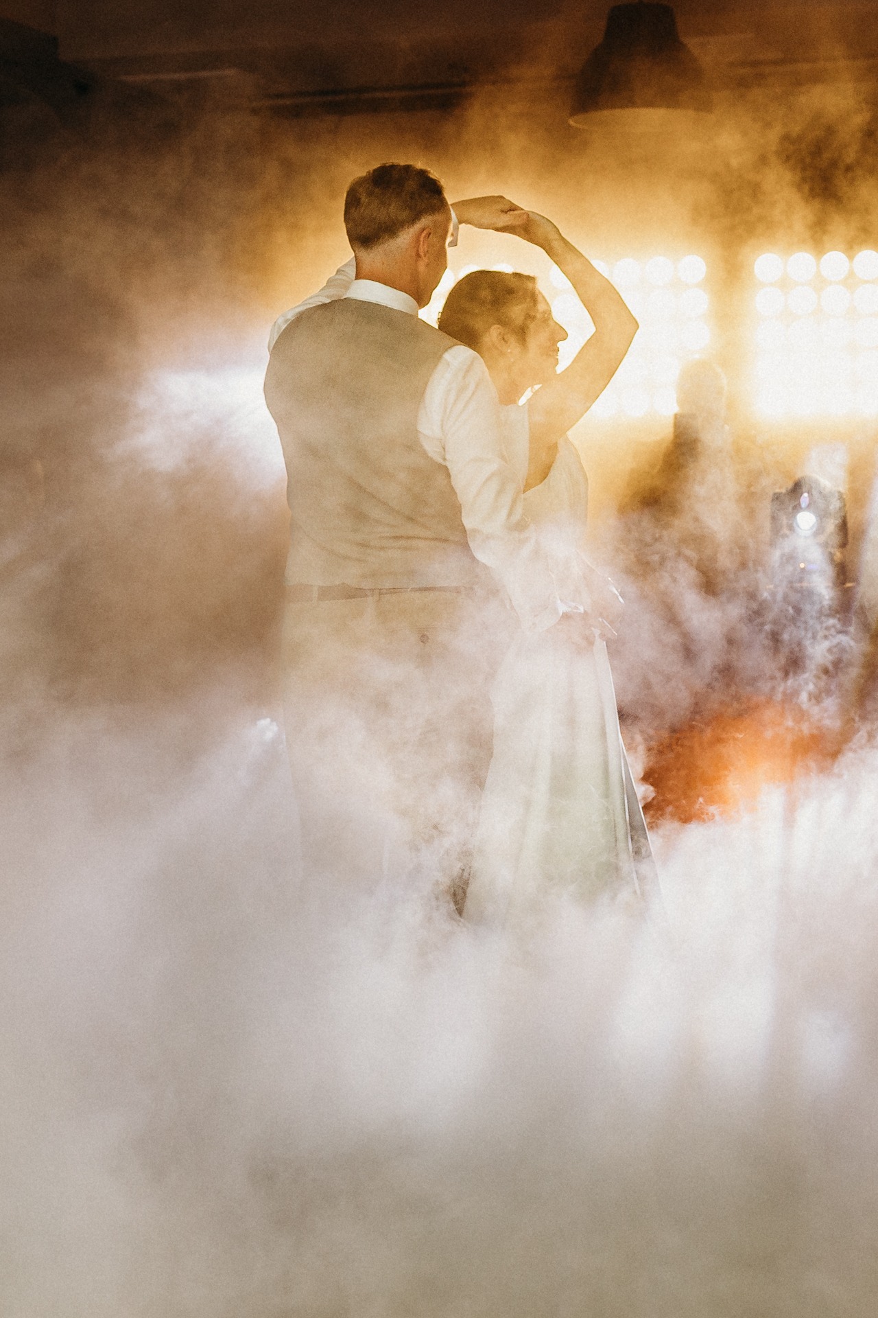 Para młoda tańczy w dymie na tle świateł scenicznych. Mężczyzna w kamizelce, kobieta w białej sukni. Romantyczna atmosfera, oświetlenie podkreśla taniec.