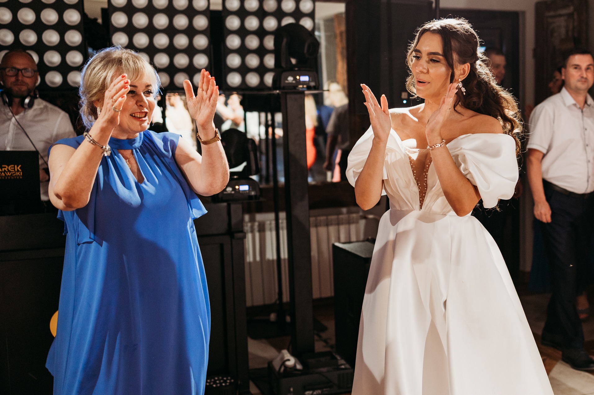 Uśmiechnięte kobiety w sukienkach klaszczą na tle oświetlenia scenicznego i DJ-a podczas wesela. Kobieta w błękitnej sukience i panna młoda w białej sukni.