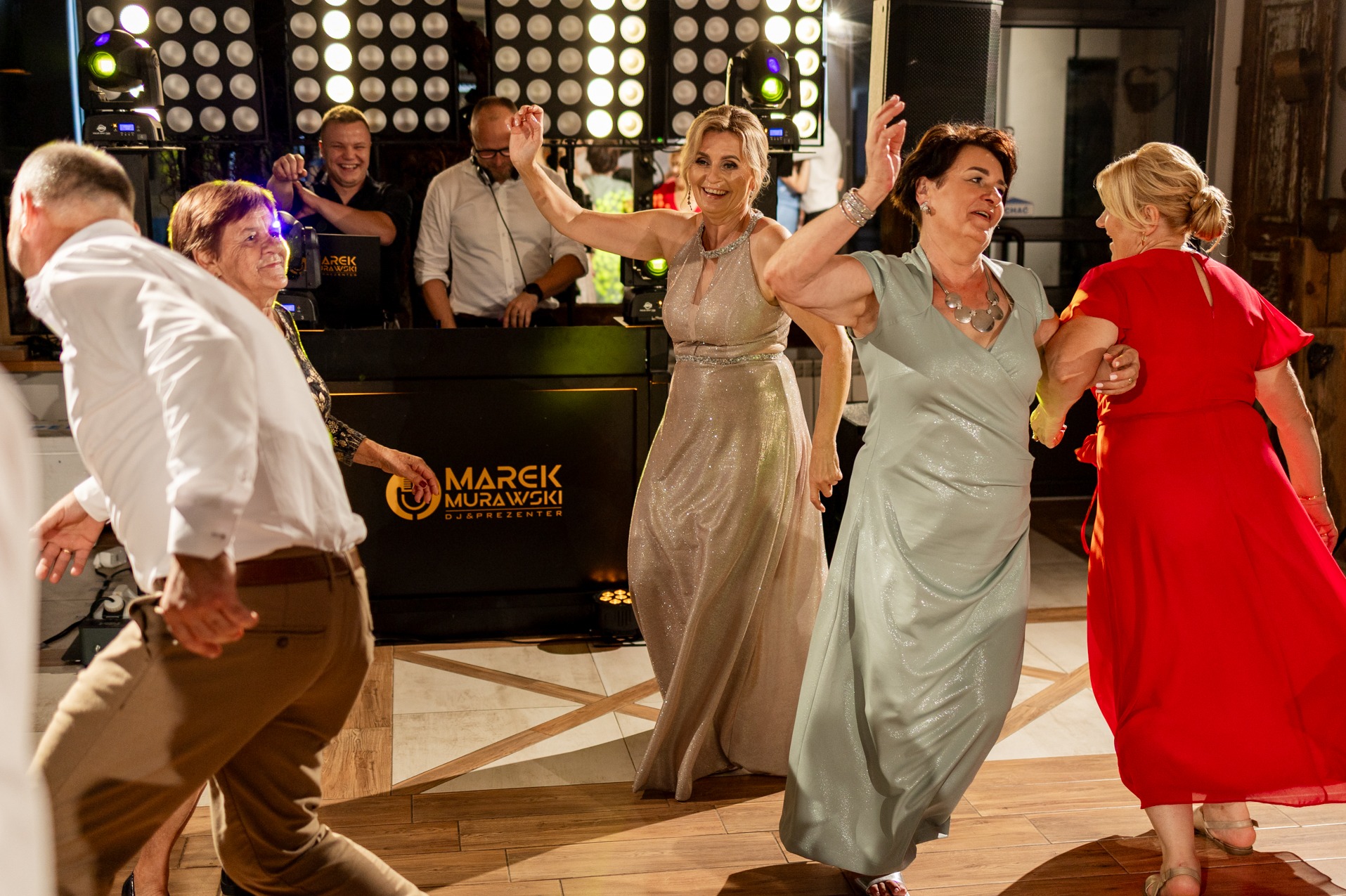 Roztańczona sala balowa z DJ-em w tle, oświetlenie punktowe, parkiet. Goście weselni w ruchu, w eleganckich strojach. Marek Murawski DJ & Prezenter.