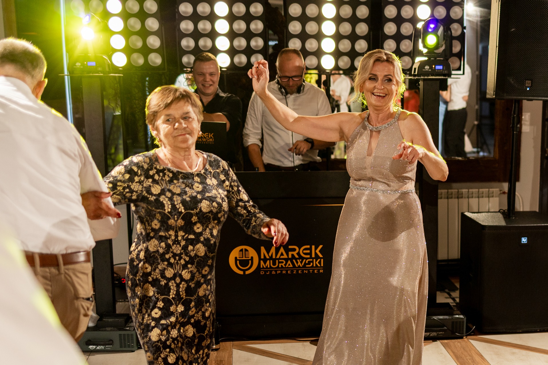 Uśmiechnięte kobiety tańczą na parkiecie, w tle DJ Marek Murawski i oświetlenie sceniczne. Radosna atmosfera imprezy.
