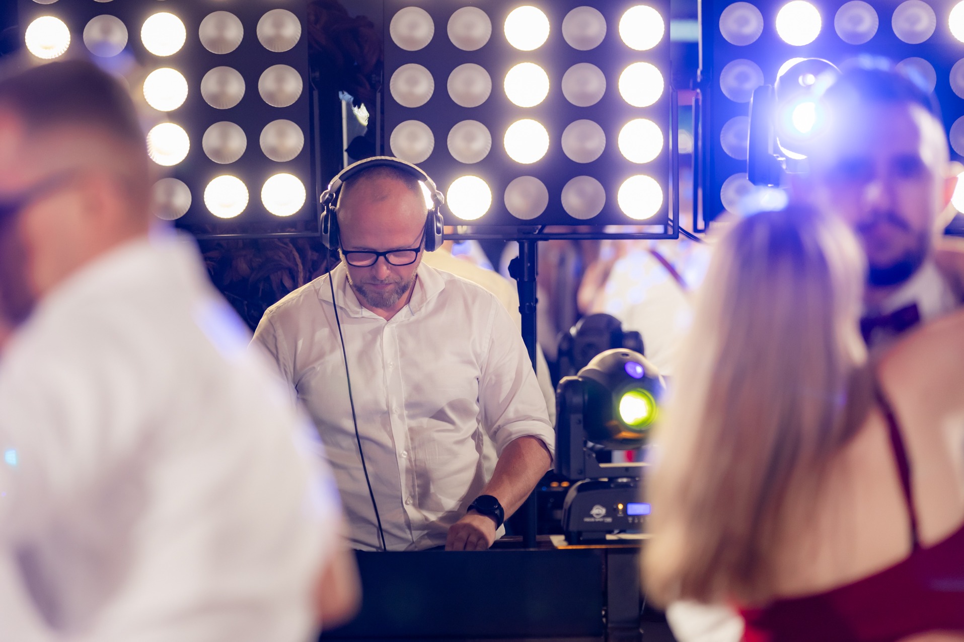 DJ w słuchawkach miksuje muzykę na tle świateł scenicznych podczas imprezy. Widoczne reflektory i osoby w tle.