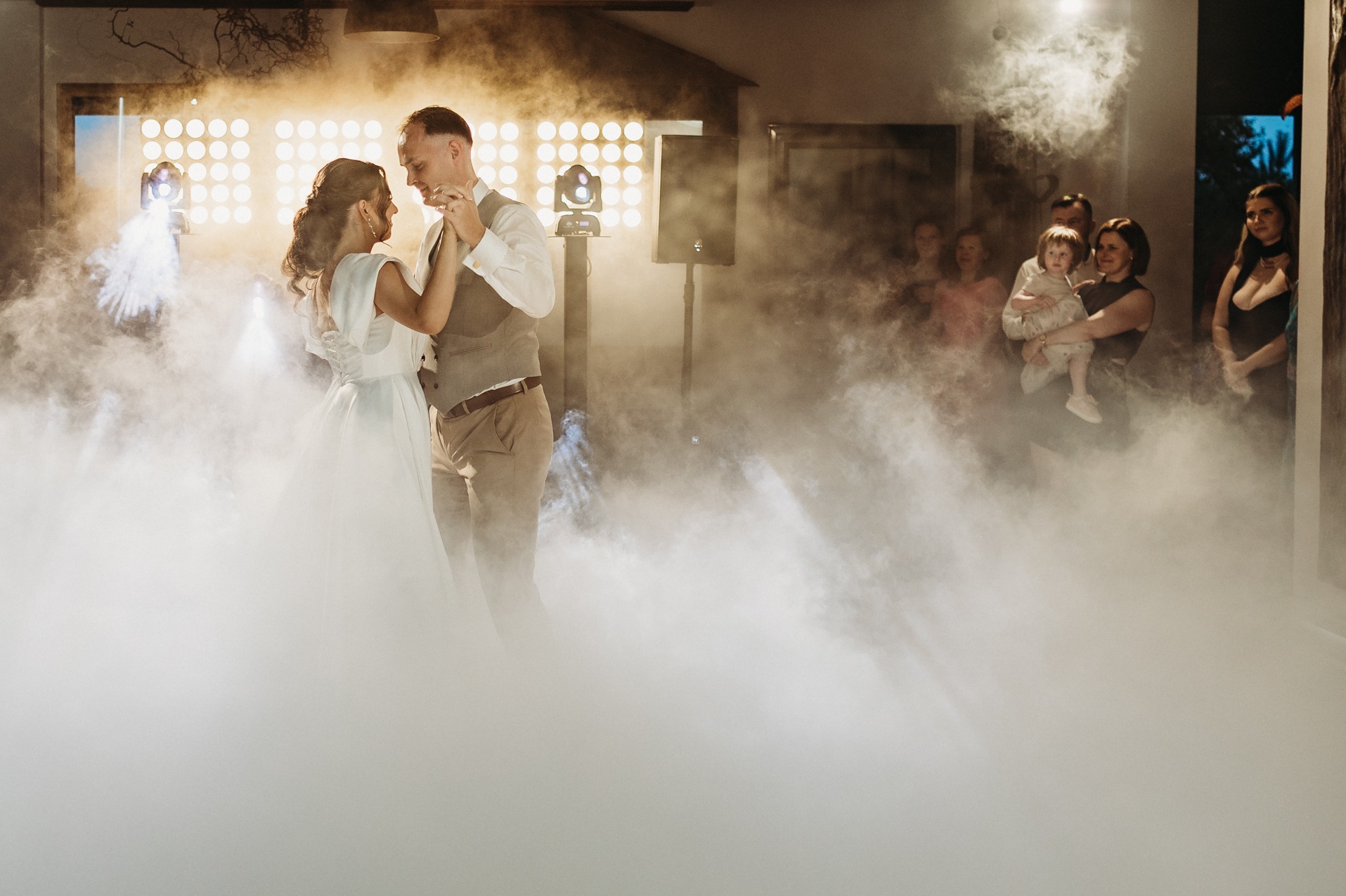 Para młoda tańczy pierwszy taniec w oparach dymu, w tle goście i oświetlenie sceniczne. Romantyczna atmosfera weselna z efektem specjalnym.